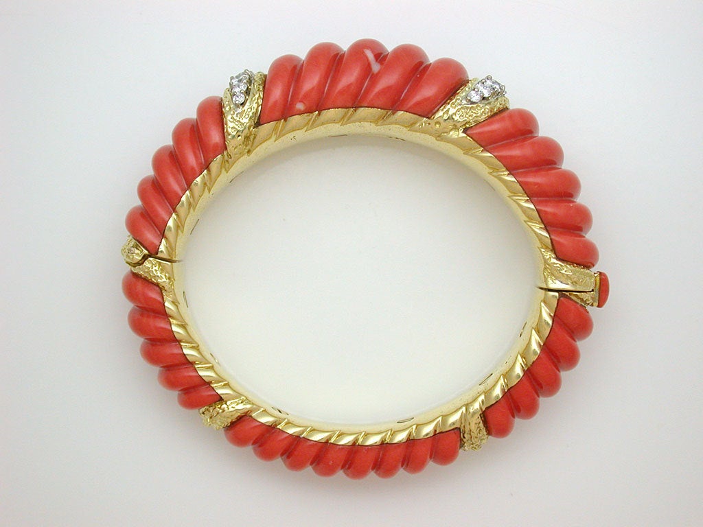 A spectacular hinged bangle, oval profile, built from carved, tapering segments of deep orange coral, accented with textured yellow gold and diamond elements on top and simpler textured yellow gold elements beneath, push button clasp and safety
