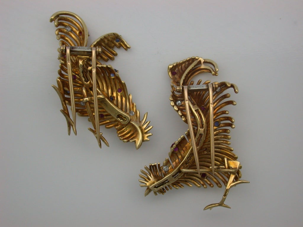 Designed like scrolling feathers, these brooches upon further inspection are actually stylized chickens, one male and one female, mounted in 18kt yellow gold and accented with circular-cut rubies, sapphires and diamonds, with French assay marks for