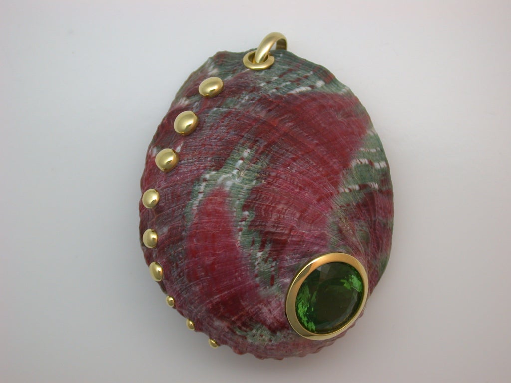 The large, domed natural shell colored raspberry pink with slate blue/grey markings, centering a fancy-cut circular green tourmaline weighing approximately 19 carats total, enhanced down one side with a swirling row of polished yellow gold buttons,