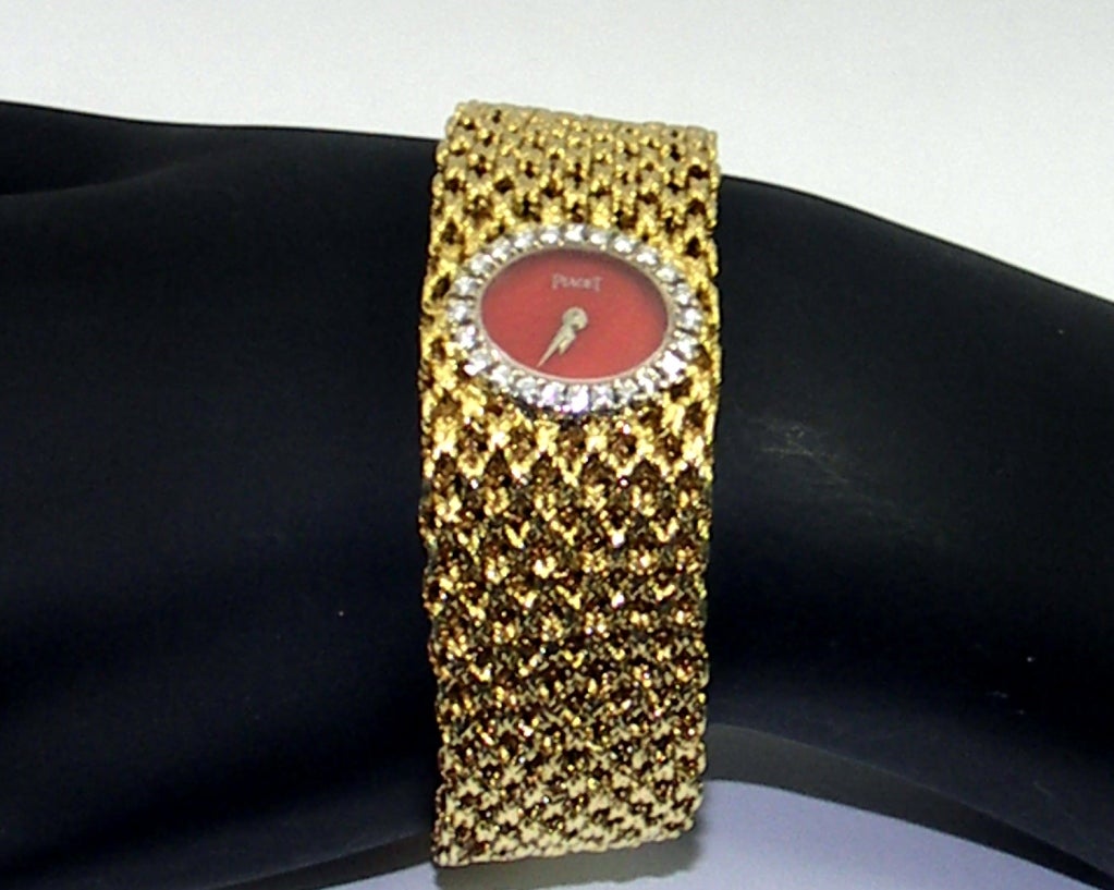 A lady's Piaget wristwatch crafted in 18k yellow gold, featuring a woven bracelet measuring 7/8 of an inch across at the dial, and tapering down to a half an inch at the buckle. The oval dial features a slice of coral. Around the dial, are 24