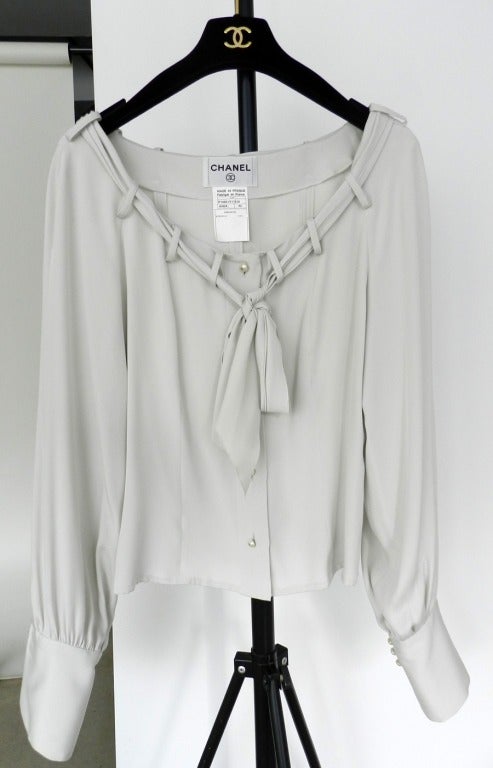 Chanel light dove grey silk blouse with sash at neckline. Excellent previously owned condition. Domed pearl buttons with silvertone CC and fold-up french cuffs.  2002 fall. Tagged IT42 / FR 38 / USA 6. Actual garment bust is 36