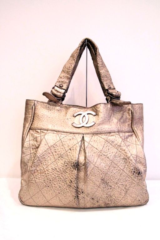 This CHANEL Le Marais Large Tote Bag is made of distressed light gold leather with antiqued pewtertone hardware. Closes with a magnetic leather strap closure on top of the bag. Double leather handles. Grey quilted fabric lining. Interior of the bag