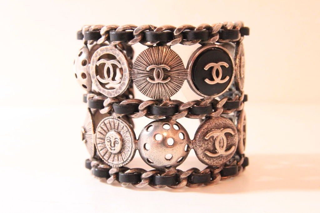 This CHANEL Pewter Tone Cuff Bracelet is both rare and collectible.  It features a black leather laced classic chain design together with 14 individual button rounds, several of which include the CHANEL “CC”.  Stamped “CHANEL” on the inside of cuff.