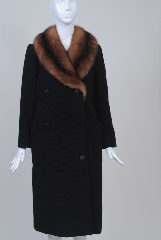 A harbinger of the silhouettes to come in the 1960s, this coat was designed by featured designer Norman Norell c. 1958, shortly before he began his namesake firm in 1960. The double-breasted, straight coat is constructed of a ribbed black wool, has