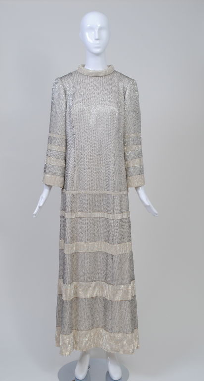 A column of shimmery brilliance, this gown is a show stopper. Composed of vertical rows of silver bugle beads with graduating bands of rhinestones on the lower half of the body,  wide sleeves, and roll collar. Back zipper. The ground is a ribbed