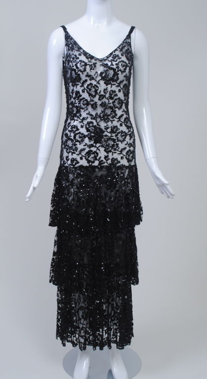 A great evening look, c. 1930, in black floral lace outlined in micro sequins. The tank-shaped dress is fitted through the body and has three deep tiers below the hips, ending at ankle length. Narrow straps with rows of the sequins. A few minor