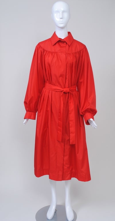 Pauline Trigère lightweight raincoat in her favorite color, red, fashioned of a synthetic silky fabric. The full coat has a pointed shirt collar and a contoured yoke from which the gathered fabric of the sleeves and body falls to mid-calf length.