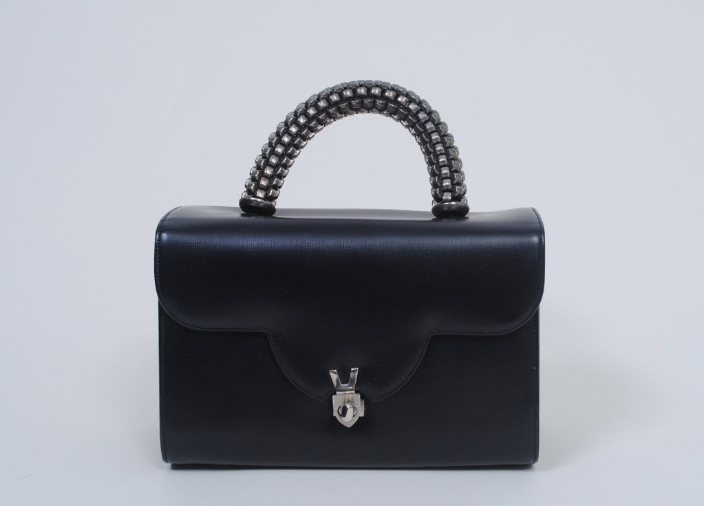 The perfect '60s handbag with hard-to-find silver trim! Black leather in a box shape with scalloped flap and a unique loop handle of cylindrical chain in burnished silver. Turn clasp and protective feet in silver metal. Open and zippered side