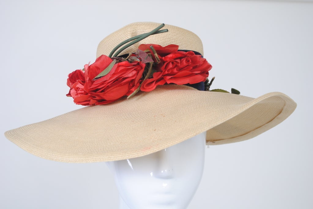 Wide-brimmed natural straw hat w/wide navy velvet band and red fabric roses. Small high crown perches on top of head, does not pull down, lined in taffeta. Wide brim, 5 1/2 inches at front tapering to 4 