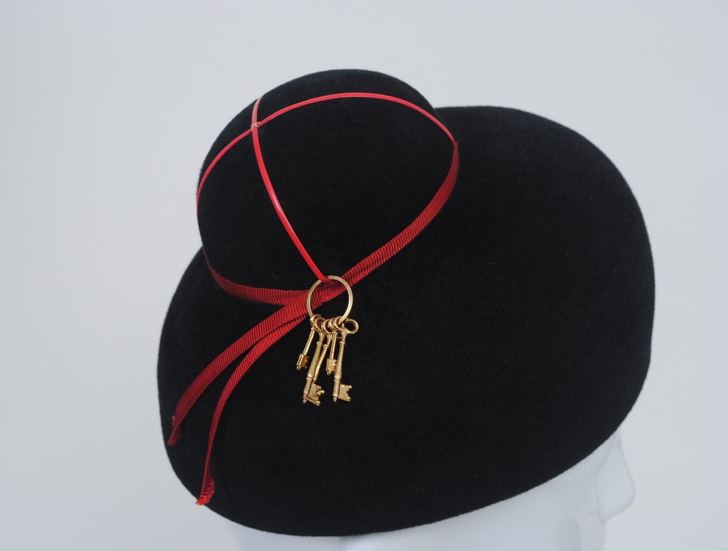Adolfo 1960s black wool felt hat in a double dome shape, the top dome like a chignon crisscrossed with red cord and wrapped around its base in narrow red ribbon with tails. A set of old fashioned keys dangle from the trim in an unusual and whimsical