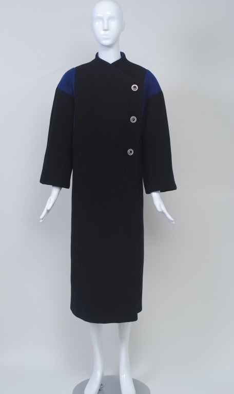 Manteau Pauline Trigére des années 1980 en laine noire avec inserts triangulaires bleu roi aux épaules et fermeture asymétrique côté gauche avec boutons bobine en métal argenté. Manches larges et col droit et décolleté en V qui suit la courbe du cou.