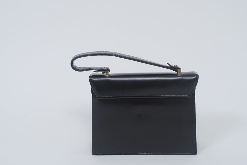 Quintessential 1960s structured black leather handbag features an unusual exterior oval covered mirror highlighted by the curve of envelope closure. Accordion body, single handle and brass hardware. The interior, lined in buff suede, is divided in