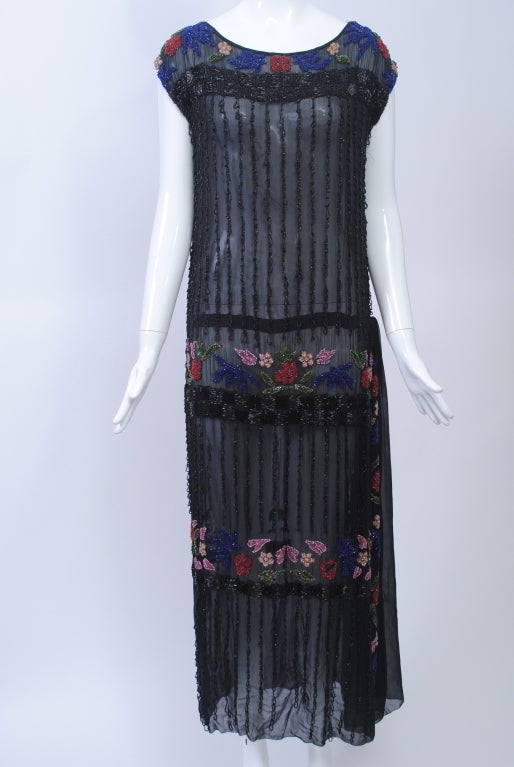 Art Deco evening chemise distinguished by bands of multi-colored floral beading bordered by a checkerboard pattern in black beads. The single-layer black silk chiffon dress is further embellished with vertical rows of looped beading with tiny black