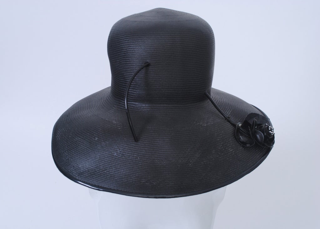 1960s dramatic black hat in fine straw by Frank Olive. High crown and wide, graduated curved brim trimmed in black patent. Accented with a black patent flower on brim whose stem arcs through brim and crown. Interior measurement 22