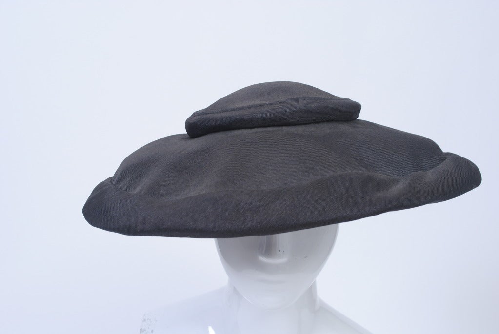 Mr. John, one of America's great milliners, was known for his classic, unembellished shapes, working under his own label from the late 1940s until 1970. Here,  a picture hat in black woven fabric with a bit of a luster, features a flat crown and a