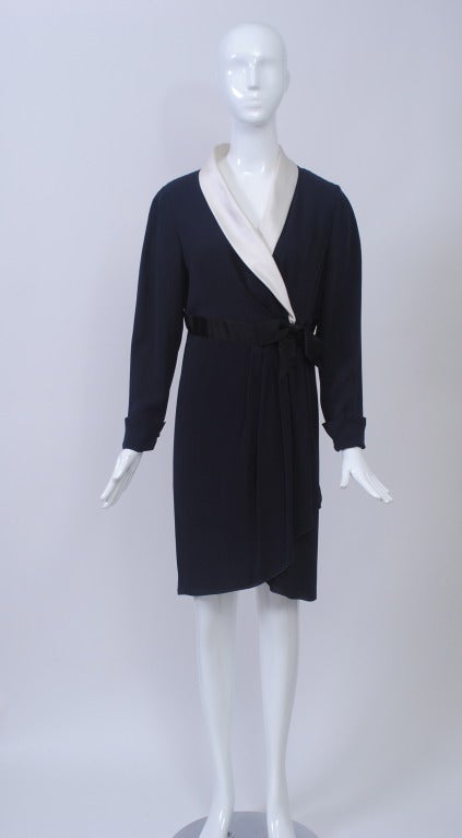 This Chanel navy silk wrap dress features a white satin shawl collar and black grosgrain waist band with bow above the waist. The skirt curves up in front over the underlying layer. The sleeves are long with a turned-back cuff and button closure.