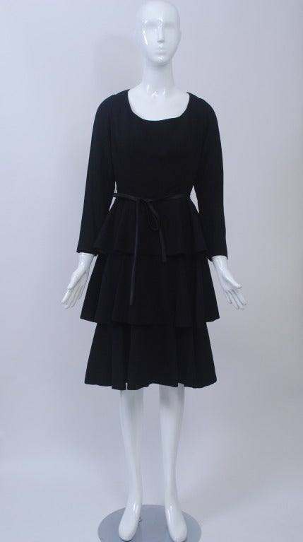 1960s dress in black wool with fitted, scoop-neck bodice and triple-tier flounce skirt. Narrow satin belt. Lined in black taffeta. Back zipper. Size S.