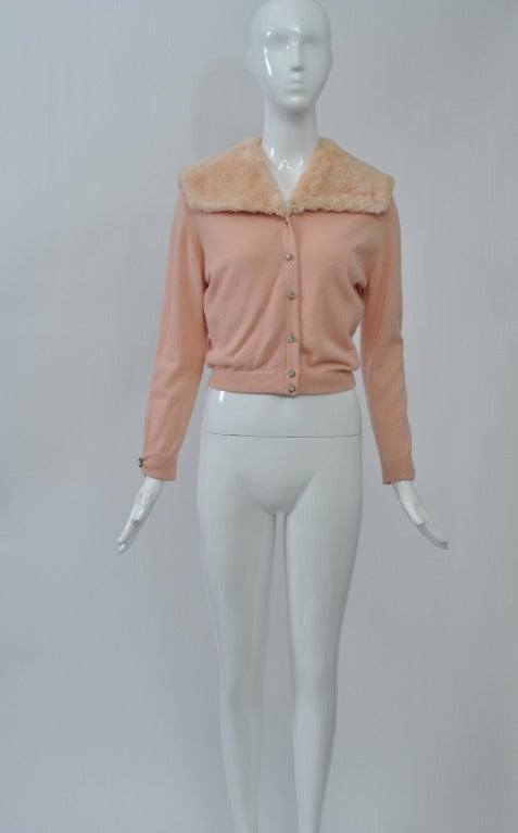An unusual example of the fur-trimmed cashmere cardigans from the 1950s and '60s, this sweater is in pale pink and is trimmed with a sailor-style collar in matching sheared beaver. Rhinestone buttons down front and at wrists. Body lined.