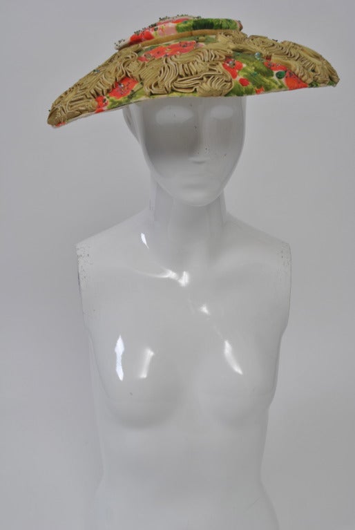 Wide brimmed and with a flat crown, this beautiful floral print hat is embellished with soutache and beads. The fabric has a beige ground with coral and green flowers and foliage. Around the crown is a narrow pale green velvet ribbon tied with a bow