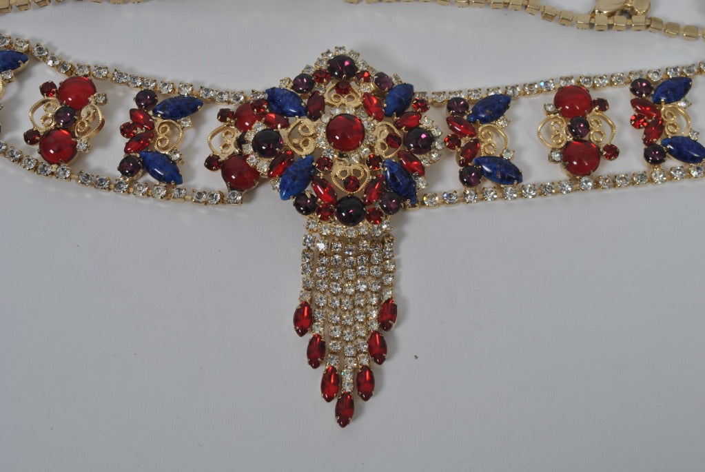 Gilt metal belt accented with red and blue stone and bordered in clear rhinestones. At center is a matching stone medallion from which hang 5 strands of rhinestones, each terminating in a marquise ruby stone. Can fit up to a 30