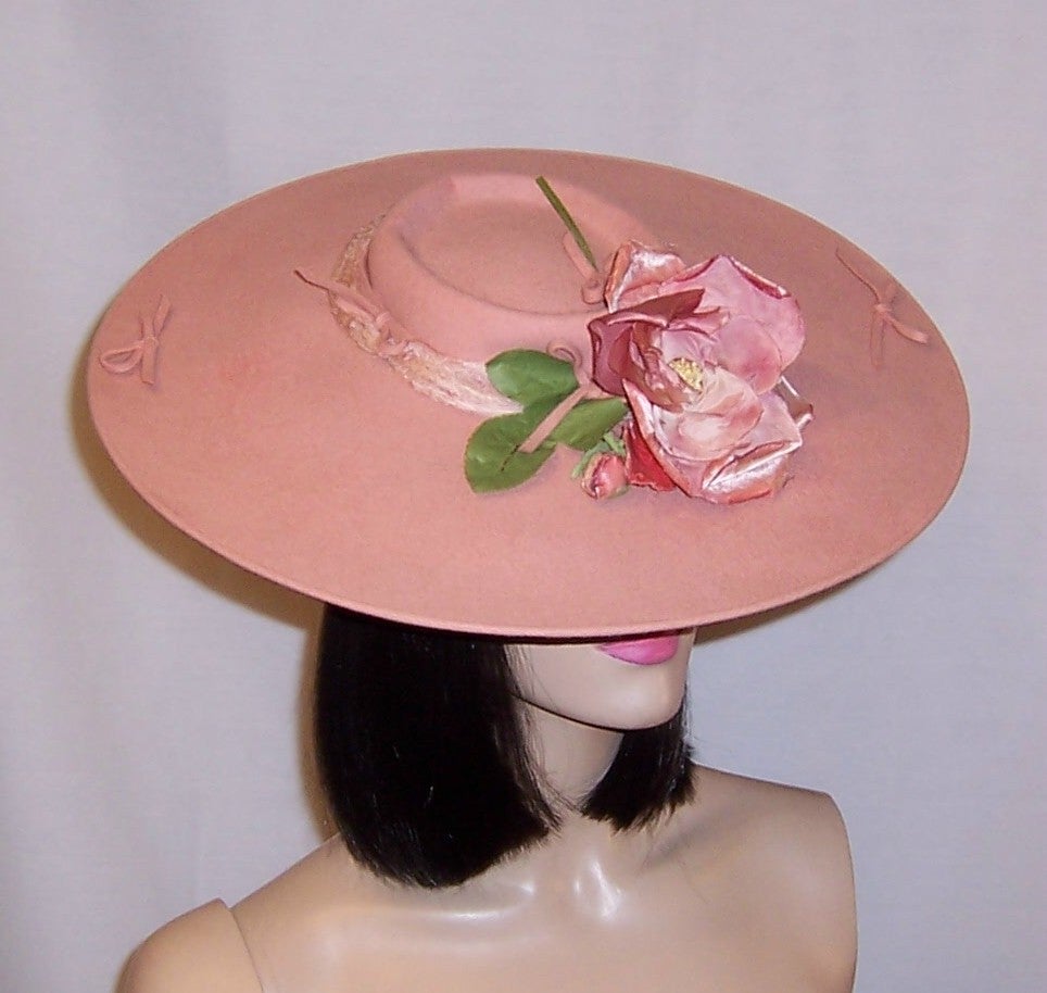 This is a delightful Depression era, late 1930's-early 1940's, pink wool felt platter hat with a single rose blossom at the front and center of its wide brim and shallow crown.  Since it has been designed to  sit on top of one's head, it will