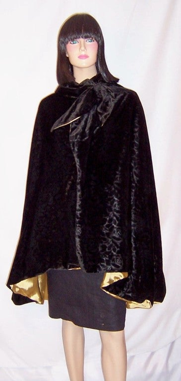 This is a simple and elegant Art Deco black velvet cape with a textured rosette pattern lined in a soft gold charmeuse.  The collar has a snap and two hooks and eyes for closure which terminates in a floppy bow. It is in excellent vintage condition