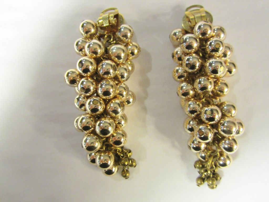 This is a striking and fanciful pair of large, clip-on, dangling earrings resembling clusters of grapes.  Each earring is constructed of many bright gold beads of uniform size attached to a chain to allow for movement. Each is then terminated with