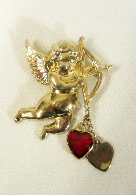 Offered for sale is this adorable gold-toned Cupid brooch holding a bow and arrow with two hearts dangling from its bow. The brooch is in excellent vintage condition and is stamped 