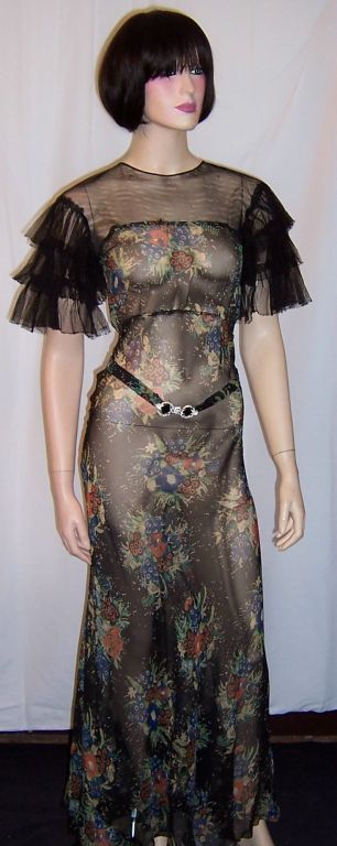 This is a dramatic 1930's, bias cut, floral printed evening gown on a black background with a detachable belt with an Art Deco buckle. The bodice and ruffled sleeves are made of fine black netting. There is a one button closure at the back of the