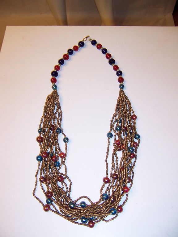 This is an unusual and attractive multi-strand necklace comprised of tiny gold seed beads interspersed with iridescent turquoise and red glass beads which hang from two strands of cobalt blue and red beads separated by smaller gold brass beads. It