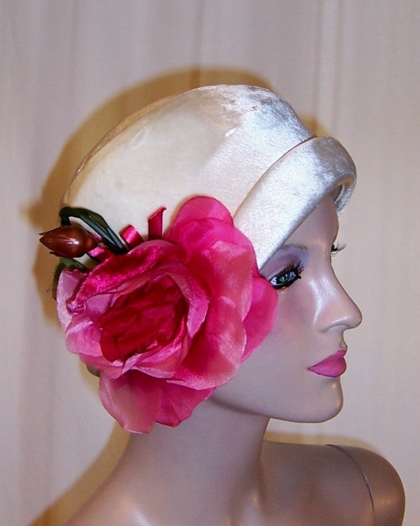 This is a handsome white silk velvet cloche dramatically embellished with an oversized deep pink silk rose blossom and bud.  The interior is lined in white fabric and is in excellent condition. Its circumference measures 22