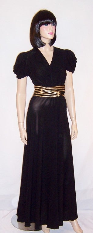 This is a fabulously sleek and sensual 1940's black crepe floor length gown with an attached gold braided striped belt which has been tied around the back of the gown and is terminated with silk tassels. The gown has a flattering V-neckline, poofy