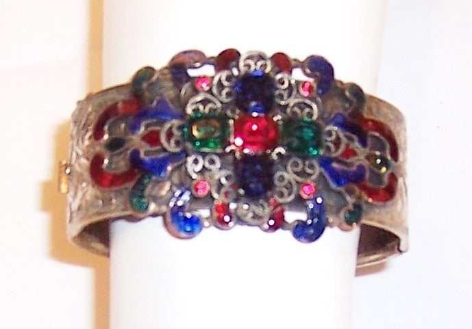 This is a gorgeous, gold-toned hinged bangle bracelet with a safety chain, lovely engraving work, filigree, enamelwork, and red, blue, and green emerald cut rhinestones. The bracelet is in very good vintage condition with the exception being that
