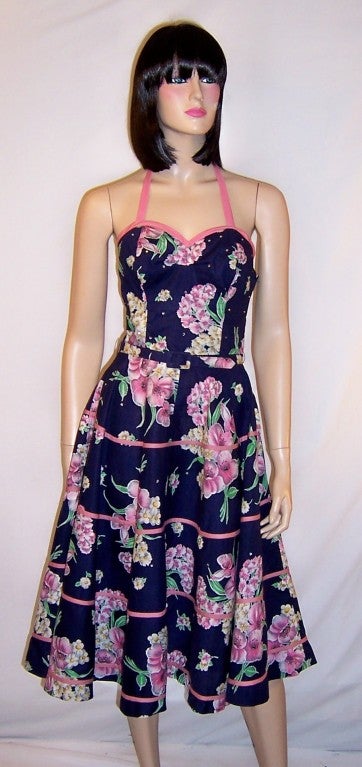 This is a striking and fanciful 50's halter-neck sundress with large sprays of pink and white flowers against a navy background. The sweetheart neckline of the bodice is trimmed with a strip of pink fabric and another strip of pink fabric rounds the