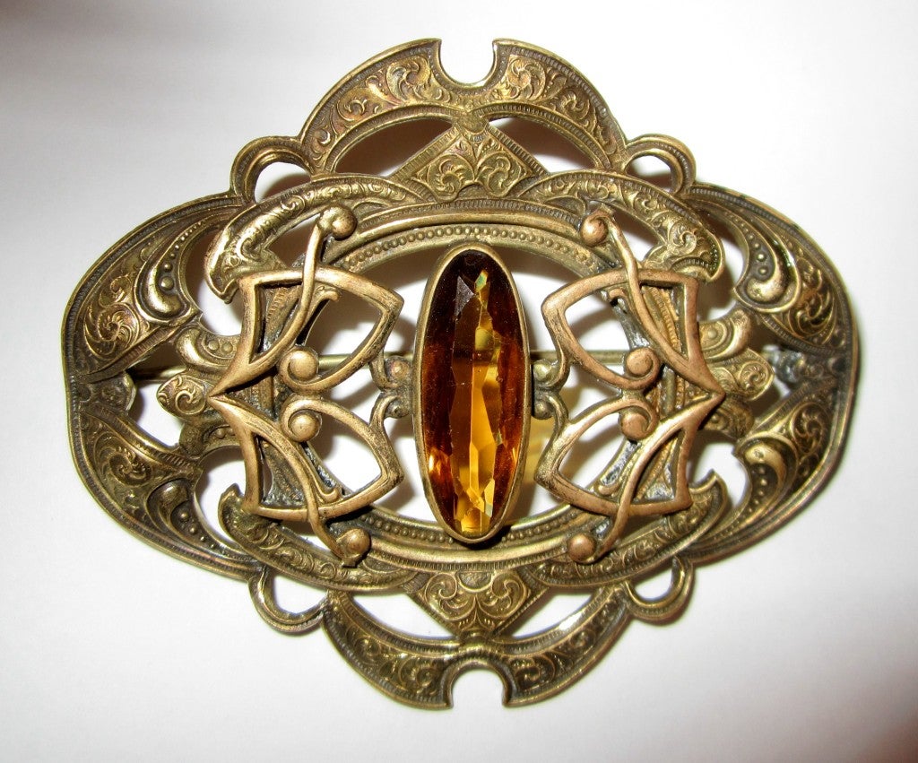 This is a large and beautifully detailed Victorian sash pin in brass with an elongated oval-shaped, cut amber-colored glass stone.  The brooch measures 3
