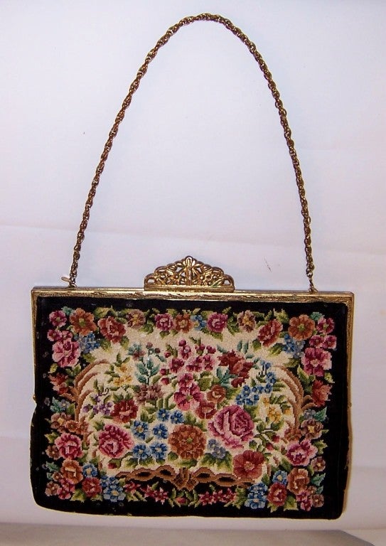 This is a 1950's petit point evening bag with a floral design in hues of red, blue, ochre, and green on a black background. The bag has a gold-toned frame and a pierced clasp, measures 7 1/2