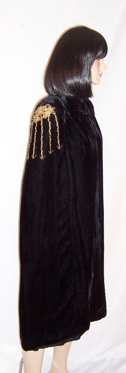 This is a luxurious black silk velvet cloak or cape with a one button closure at its neckline, a slit at either waist/hip area for one's arms, slightly padded shoulders, and gold metallic embroidery with tiny brass studs embellishing each shoulder.