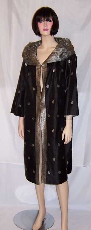 This is a handsome, custom-made evening coat, of the 1960's vintage, made of the finest black silk and embellished with silver brocaded trim and full collar which stands up and frames the face. It had been manufactured by Royal India and custom-made
