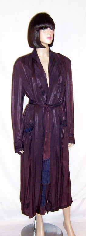 Offered for sale in this original, men's Art Deco, striped smoking/lounging robe in a deep aubergine color.  The robe has a breast pocket and a pocket at each hip.  It has a belt with long fringe for closure and is fully lined. It is in excellent
