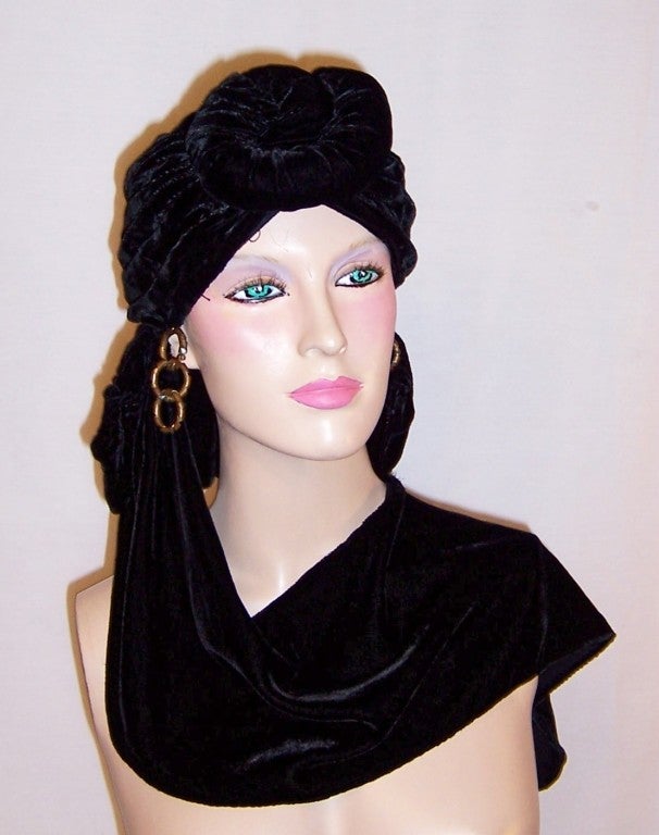 This is a dramatic and unusual 1940's black velvet knotted turban with its own attached drape which can be worn over the shoulder.  The turban has a huge knot in the front and center, attached brass hooped earrings and either side, a wired