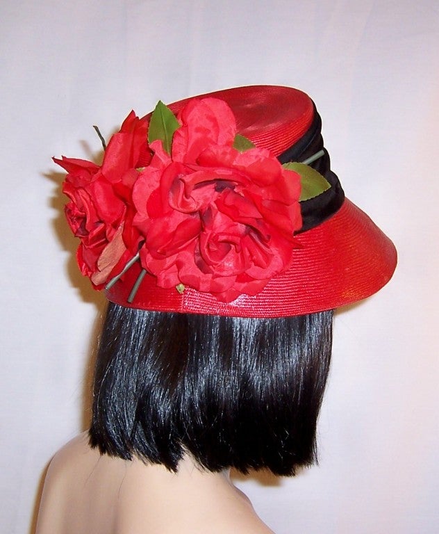This is an elegant red straw chapeau whose crown is encircled with a wide black band and embellished with two large red silk rose blossoms at its back. It is of the 1960's vintage and its label is in tact and reads Christian Dior Paris-New York.  It