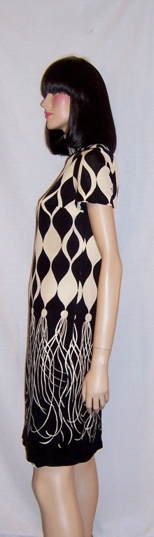 This is a striking black and white Mod-couture shift dress made in Hong Kong by S. Andrews. The dress has short sleeves, a long back zipper for closure, an overall black and white stylized Harlequin design pattern, and white fringe done in a