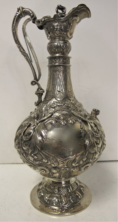 A magnificent large antique sterling silver wine jug of elegant shaped form on pedestal foot. The hinged lid, with ornate flower finial, opens by squeezing the handle mechanism. Beautifully chased with flowers, swirls and leaves, the jug has two