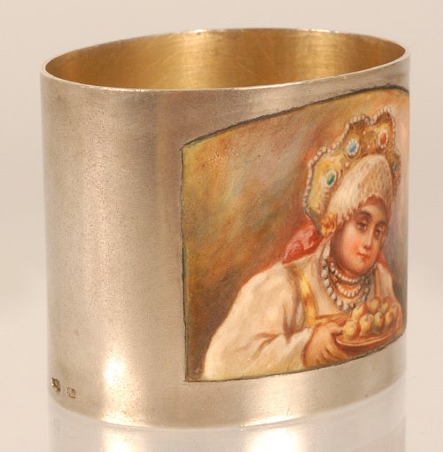 A Russian silver and parcel gilt napkin ring, Peter Mulikov, Moscow, 1908-1917. The oval ring enameled with an en plein depiction of a traditionally dressed young woman presenting a plate of sweets. Dimensions: 1 7/8
