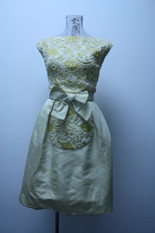 This dress reminds me of Doris Day. It is a pale colour of creme yellow with lemon accents in its brocade bodice.The brocade shell is accented with Austrian crystals. It has a big satin bow at the front. It sports a boat neckline and a lower scoop