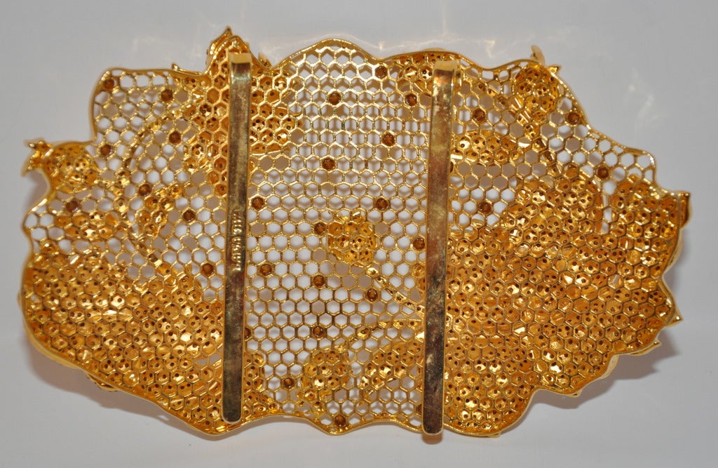 Die extragroße, florale Gürtelschnalle von Badia Taxi ist mit Strasssteinen in Bronze und Kanariengelb sowie mit rubinroten Strasssteinen verziert. Die floralen Motive werden auf verschiedene Ebenen angehoben, um einen 3-D-Effekt zu erzielen. 
  