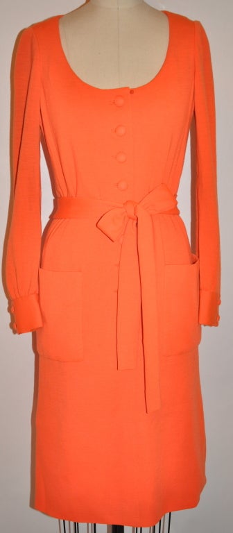 This wonderful timeless Iconic Norman Norell fully lined dress is in a neon tangerine hue. The lining is in silk of the same hue. The dress has a attached self-tie belt which measures 79 inches x 2 inches. There are two patch pockets on front.
   As