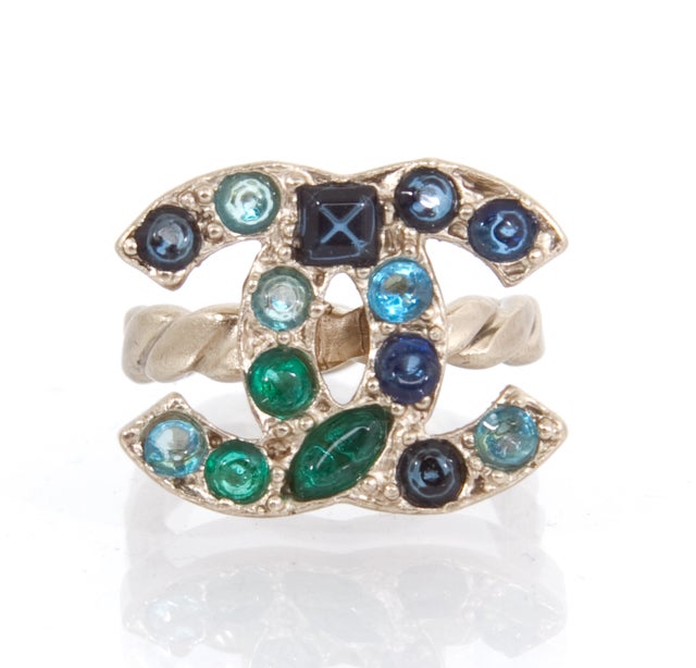 This is an authentic CHANEL Multicolor Gem Stone CC Logo Ring. It is done in silver toned hardware and features a CC logo face. The ring portion is done in a twist design and within the logo are lovely blue and green jewels. It's just stunning.