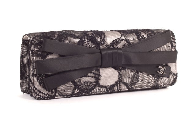 This is an authentic CHANEL Black Lace and Satin Clutch Bag Purse. This elegant piece is done in a soft silver color with a black lace overlay. There is a large bow on the front exterior that includes the interlocking CC logo. The bag closes with a
