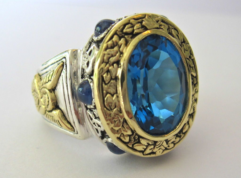 6 carat Blue Topaz set with 2.7 carats of multi-colored Sapphires, .925 Sterling Silver & 18K Yellow Gold. (9.0 grams of 18K Yellow Gold.) The “Aleluya” ring features hand carved, intricate religious details, depicting Christian imagery. 
Around